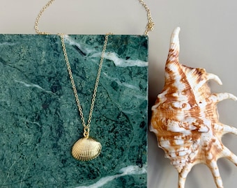 May include: A gold-colored seashell pendant necklace with a delicate chain. The pendant is a detailed replica of a seashell. The necklace is displayed on a green marble block, next to a large, brown and white seashell.