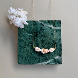 Peut inclure: Un collier à chaîne dorée orné de quatre perles en pierre blanches et orangées. Le collier est présenté sur un bloc de marbre vert, avec un morceau de corail blanc en arrière-plan.