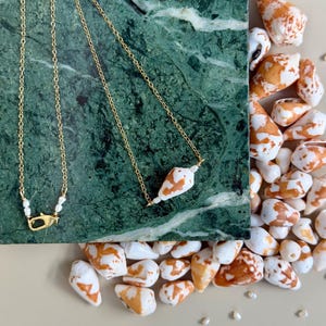 May include: Two gold necklaces with seashell pendants rest on a green marble surface. One necklace features a pearl clasp. Scattered around are white and orange seashells, creating a coastal theme.