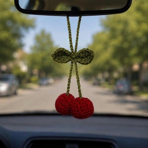 May include: A handmade crochet car accessory featuring two round, textured red cherries attached to long green stems with two small green leaves at the top, designed to hang from a rearview mirror with a looped green yarn cord.