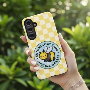 Puede incluir: Funda de teléfono a cuadros amarillos y blancos con una abeja de dibujos animados y el texto "ENTOMOLOGY CLUB LIFETIME MEMBER". La funda del teléfono tiene una cámara de triple lente.