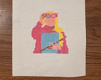 May include: Cross-stitch of a character with blonde hair, pink glasses, and a pink outfit holding a blue book and a wand. The cross-stitch is on a white fabric background, set against a wooden surface.