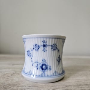 May include: A white ceramic cup with a blue floral pattern. The cup has a ribbed texture and a flared shape. The blue floral design includes flowers and leaves. The cup is on a wooden surface.