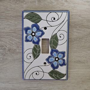 Peut inclure: Plaque de prise de courant décorative en céramique avec un motif floral. Le couvercle blanc a une bordure bleue et est orné de fleurs bleues, de feuilles vertes et de lignes noires en spirale. Il comporte une ouverture pour un interrupteur et deux trous de vis.