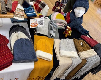 May include: A selection of winter hats, scarves, and gloves in a variety of colours and patterns. The hats include beanies and cable-knit styles, with a mannequin head wearing a blue hat and scarf. The items are arranged on a table with a white tablecloth.