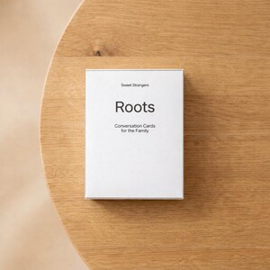 Peut inclure: Une boîte blanche de cartes de conversation "Roots" pour la famille, avec le texte "Sweet Strangers" au-dessus du titre. La boîte est posée sur une surface en bois brun clair.