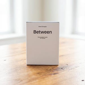 Peut inclure: Une boîte blanche de cartes de conversation intitulée "Between" avec le texte "Sweet Strangers" au-dessus et "Conversation Cards for Friends" en dessous. La boîte est posée sur une table en bois.