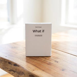 Puede incluir: Una caja blanca de tarjetas de conversación "What if" de Sweet Strangers está sobre una mesa de madera. La caja tiene el texto "Conversation Cards to Think Differently". La mesa tiene vetas de madera natural.