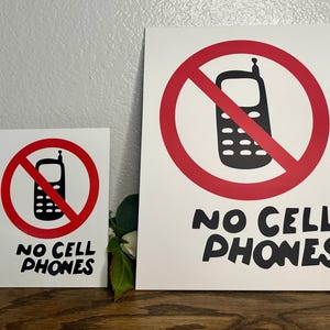 Può includere: Due cartelli rettangolari bianchi con una grafica nera di un telefono cellulare all'interno di un cerchio rosso con una barra diagonale. Sotto la grafica, il testo NO CELL PHONES è stampato in grassetto nero su entrambi i cartelli di dimensioni diverse.