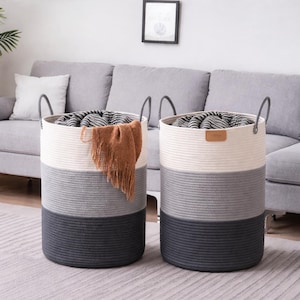 May include: Two round, three-tone laundry baskets. The top section is white, the middle is gray, and the bottom is dark gray. Each basket has two gray handles and is filled with blankets and a brown throw blanket.