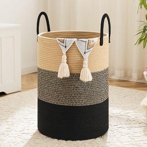 May include: A round, woven storage basket with a three-tone design. The top section is tan, the middle is black and white, and the bottom is solid black. It has black handles and decorative white tassels.