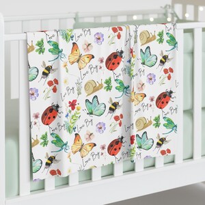 May include: White baby blanket with a colorful pattern of butterflies, ladybugs, bees, snails, flowers, and dragonflies. The words "Love Bug" are written throughout the design. The blanket is draped over a white crib.