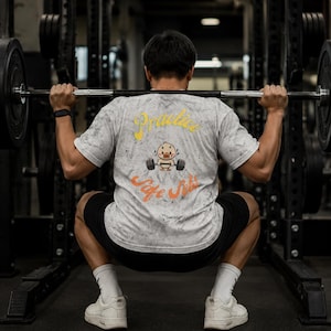 Könnte beinhalten: Eine Person, die in einem Fitnessstudio Kniebeugen mit einer Langhantel macht, trägt ein weißes und graues marmoriertes T-Shirt mit dem Text "Practice" und einer Cartoon-Ente, die Gewichte hebt. Die Person trägt schwarze Shorts, weiße Socken und weiße Turnschuhe.