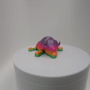May include: A colorful, 3D-printed figurine of a stylized animal, possibly a bull or rhinoceros. The figure is primarily purple, pink, orange, and green. It is resting on a white, cylindrical surface.
