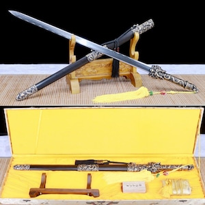 May include: Two swords with ornate dragon-themed handles and scabbards are displayed on a wooden stand. One sword is unsheathed, revealing a silver blade. A yellow tassel hangs from the hilt. The second image shows the sword in a yellow lined box.