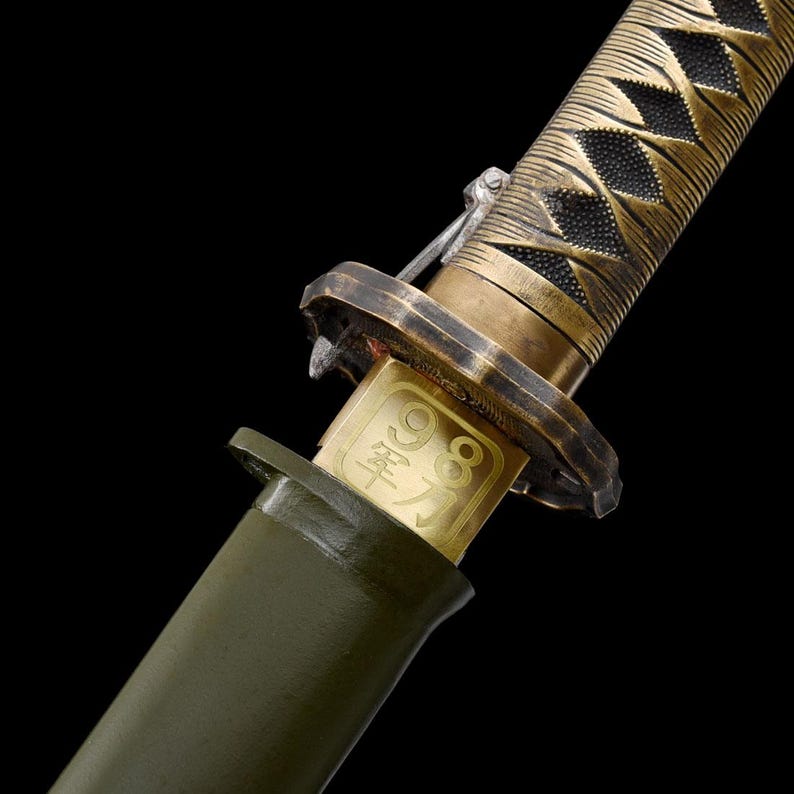 May include: Close-up of a katana sword with a gold-colored hilt and a dark green handle. The hilt features a diamond pattern. A gold plate with Japanese characters is visible near the handle.