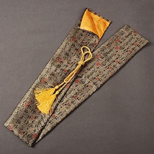 May include: An open, rectangular case with a patterned exterior in shades of gray, red, and black. The interior is a bright orange. A gold-colored tassel and cord are attached to the case. The case is likely designed to hold a long, thin object.