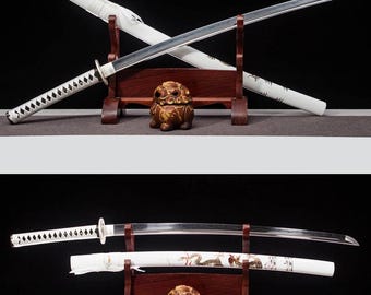 May include: Two Japanese katana swords with white scabbards and handles, displayed on a wooden stand. The blades are silver, and the handles have a white, diamond-shaped pattern. A small, brown decorative lion statue sits below the swords.