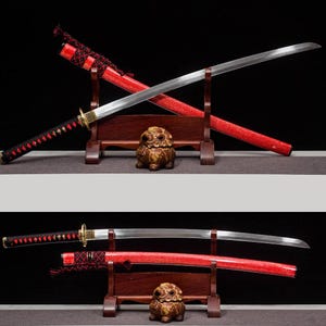 May include: Two Japanese katana swords with red scabbards and black handles are displayed on wooden stands. The blades are silver, and the handles are wrapped with black cord and red accents. A small, ornate, brown statue sits between the stands.