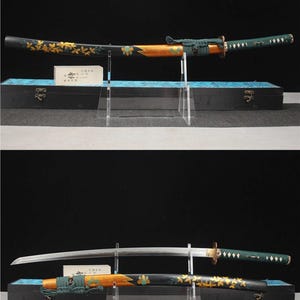 Op de afbeelding: Een Japanse katana-zwaard met een zwarte schede versierd met gouden bladmotieven. Het handvat is gewikkeld in groene stof. Het zwaard wordt tentoongesteld op een transparante standaard, met een zwarte doos op de achtergrond.
