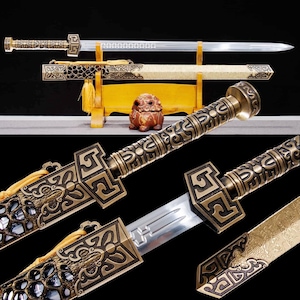 May include: An ornate sword with a bronze-colored handle and scabbard. The blade is silver and the scabbard features intricate designs. A small, brown decorative statue is in the background. The sword is displayed on a wooden stand.