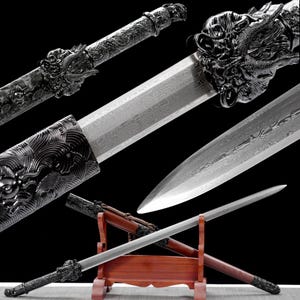 Peut inclure: Épée décorative avec une lame argentée et une garde ornée de dragons. L'épée est présentée dans un support en bois, mettant en valeur son design complexe. La lame présente un motif unique, soulignant le savoir-faire.