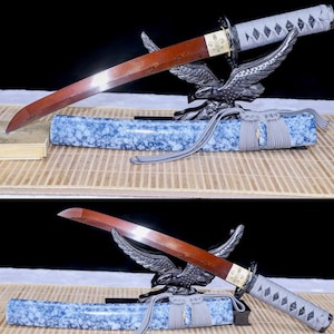 Op de afbeelding: Een Japanse tanto-mes met een roodbruin lemmet en een grijze handgreep. Het lemmet rust op een decoratieve adelaarstandaard, met daaronder een blauw-wit patroon schede. De handgreep is gewikkeld in een ruitpatroon.