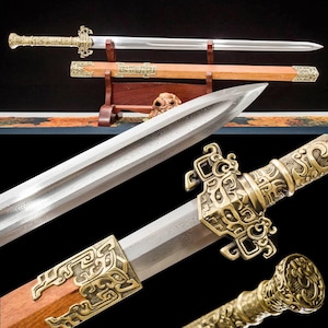 May include: A detailed image of a decorative sword with a polished steel blade and ornate gold-colored metalwork. The sword rests in a wooden scabbard with matching gold accents. The handle features intricate carvings.