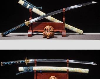 May include: Two Japanese katana swords with ornate handles and sheaths, displayed on a wooden stand. The blades are silver, and the handles are black with blue accents. The sheaths are cream-colored with a blue and gold pattern. A small, brown decorative statue sits below.