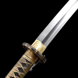 May include: Close-up of a Japanese katana sword. The blade is silver and the handle is wrapped in a gold and black pattern. The guard is gold with a decorative design. The sword is set against a black background.