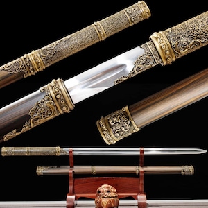 May include: An ornate sword with a polished steel blade and a wooden handle. The handle and scabbard feature intricate gold-colored metal detailing. The sword rests on a wooden stand, showcasing its craftsmanship and design.