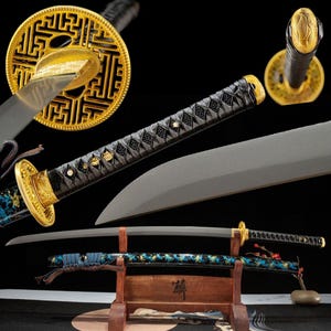 Könnte beinhalten: Ein Katana-Schwert mit einer matten grauen Klinge, einem schwarz umwickelten Griff und einem goldfarbenen runden Parierstück mit kompliziertem geometrischem Muster. Die Scheide ist schwarz mit blauen und goldenen Akzenten auf einem Holzständer.