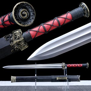 Peut inclure: Épée décorative avec une lame argentée et une poignée noire enveloppée de cordon rouge et noir. La garde et le pommeau présentent des ornements en métal doré. L'épée est présentée avec son fourreau noir.