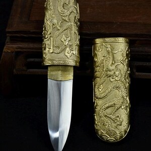 Puede incluir: Un cuchillo decorativo con una hoja plateada y una vaina y un mango dorados. La vaina y el mango están intrincadamente tallados con un diseño de dragón. El cuchillo se exhibe sobre una superficie oscura.