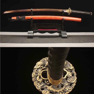Könnte beinhalten: Zwei japanische Katanas auf einem schwarzen Ständer ausgestellt. Eines hat eine braune Holzscheide und einen Griff, das andere eine rote Scheide. Die Griffe sind mit schwarzem Stoff umwickelt. Der Parier eines Katanas ist goldfarben mit einem komplizierten Design.