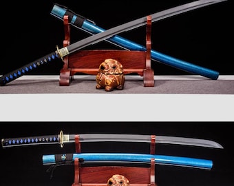 May include: Two Japanese katana swords with black and blue handles and sheaths, displayed on wooden stands. The blades are silver, and the handles are wrapped with blue diamond-shaped accents. A small, ornate brown figurine sits between the stands.