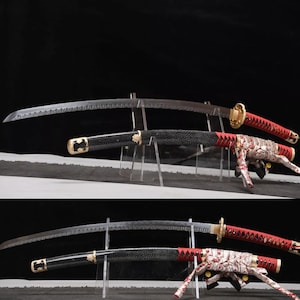 May include: Two Japanese katana swords displayed on clear stands. The blades are silver with a serrated edge. The handles are black with gold accents and red and white patterned wrapping. The swords are set against a dark background.