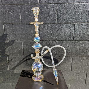 May include: A clear glass hookah with iridescent accents. The hookah has a bulbous base, a central column with two spherical sections, and a top bowl. A white hose with a decorative mouthpiece is attached.