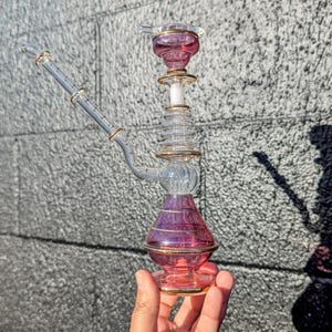 May include: A clear glass hookah with purple and gold accents. The hookah has a bulbous base, a cylindrical body, and a slender, angled stem. The design includes gold trim and a decorative top. The hookah is held in a person's hand.