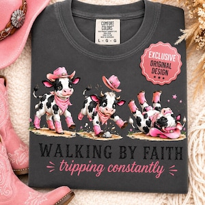 May include: A charcoal gray t-shirt featuring three cartoon cows wearing pink cowboy hats and boots. The text reads "WALKING BY FAITH tripping constantly" in black and pink lettering. A pink scalloped badge states "EXCLUSIVE ORIGINAL DESIGN".