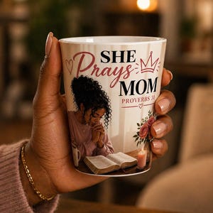 Może przedstawiać: Biały ceramiczny kubek z napisem "SHE PRAYS MOM" i "PROVERBS 31". Kubek przedstawia ilustrację kobiety modlącej się nad otwartą książką, ze świecą i kwiatami. Kubek jest trzymany w dłoni.