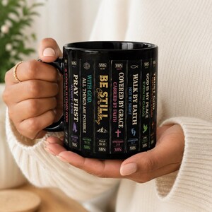May include: Black ceramic mug featuring inspirational quotes and Bible verses printed on a VHS tape design. The mug includes phrases like "Pray First" and "Trust God." A unique gift for those seeking daily inspiration.