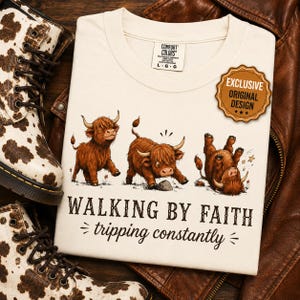 Puede incluir: Una camiseta de color crema con tres ilustraciones de ganado Highland marrón y el texto "WALKING BY FAITH tripping constantly." La camiseta incluye una etiqueta Comfort Colors y una insignia "EXCLUSIVE ORIGINAL DESIGN."