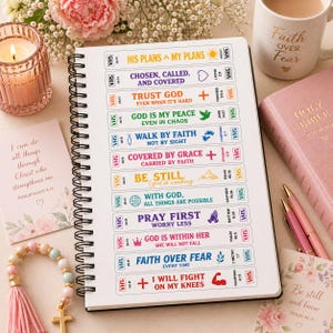 Puede incluir: Un cuaderno de espiral con citas inspiradoras y versículos bíblicos, incluyendo "Fe sobre el miedo" y "Lucharé de rodillas". El cuaderno abierto muestra texto y gráficos coloridos. Una vela encendida, una taza y una Biblia rosa también son visibles.