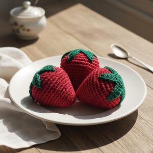 May include: Three handmade crochet strawberries with green tops rest on a white plate. The red fruit is arranged on a wooden table, with a sugar bowl and spoon in the background. The scene is lit by natural light.