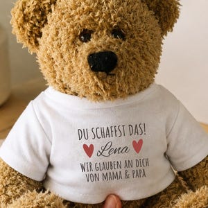 Könnte beinhalten: Ein brauner Plüsch-Teddybär, der ein weißes T-Shirt trägt. Auf dem T-Shirt steht der Text "DU SCHAFFST DAS!" über dem Namen "Lena", flankiert von zwei roten Herzen, mit dem Text "WIR GLAUBEN AN DICH VON MAMA & PAPA" unter dem Namen.