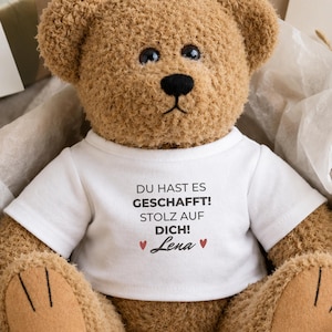 Könnte beinhalten: Ein brauner Plüsch-Teddybär trägt ein weißes T-Shirt mit dem schwarzen Aufdruck DU HAST ES GESCHAFFT! STOLZ AUF DICH! und dem Namen Lena, flankiert von zwei kleinen roten Herzen. Der Bär hat dunkle Augen, eine schwarze Nase und beige Filzpfoten.