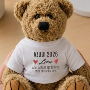 Könnte beinhalten: Ein brauner Plüsch-Teddybär in einem weißen T-Shirt. Das Shirt trägt den Text AZUBI 2026, den Namen Leon zwischen zwei roten Herzen und den deutschen Satz Aller Anfang ist schwer, aber du rockst das in schwarzer Schrift.
