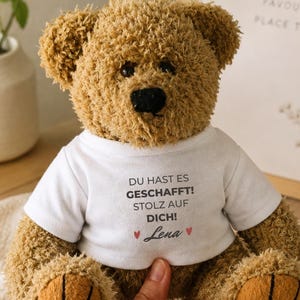 Può includere: Un orsacchiotto di peluche marrone che indossa una maglietta bianca con il testo stampato in nero DU HAST ES GESCHAFFT! STOLZ AUF DICH! e il nome Lena in corsivo, affiancato da due piccoli cuori rossi, con occhi scuri e naso nero.