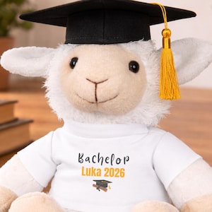 Può includere: Un peluche a forma di agnello con un cappello di laurea nero e un nappa dorata. L'agnello indossa una maglietta bianca con la scritta "Bachelor Luka 2026" e un'immagine del cappello di laurea. L'agnello è su una superficie di legno.